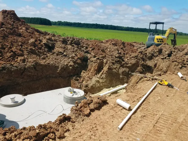 Excavation site with a trench, exposed white foundation, dirt piles, and a backhoe in the distance