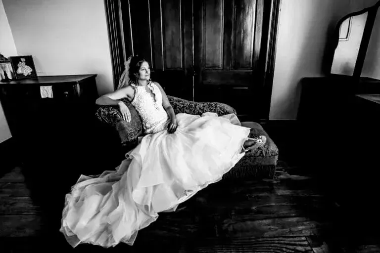 Bride in wedding dress sits on a chaise lounge in a room with dark wood accents
