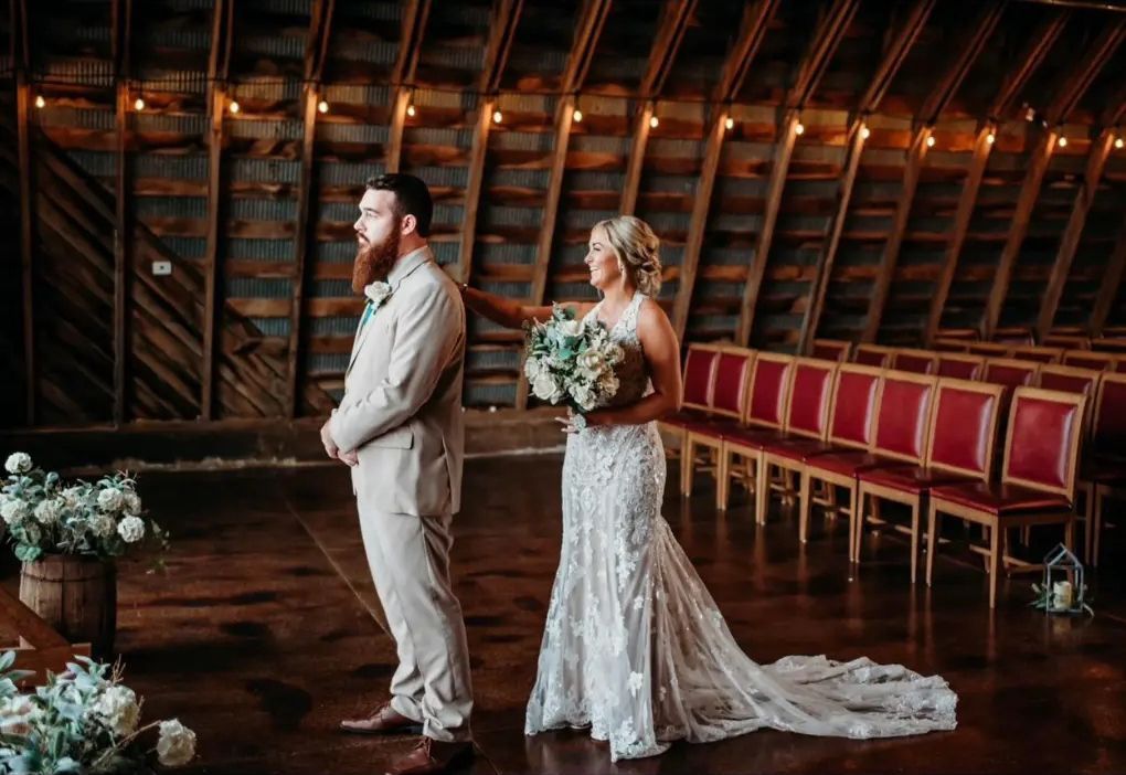 Bride touches groom's shoulder in rustic barn wedding. She wears a lace gown, he a tan suit.