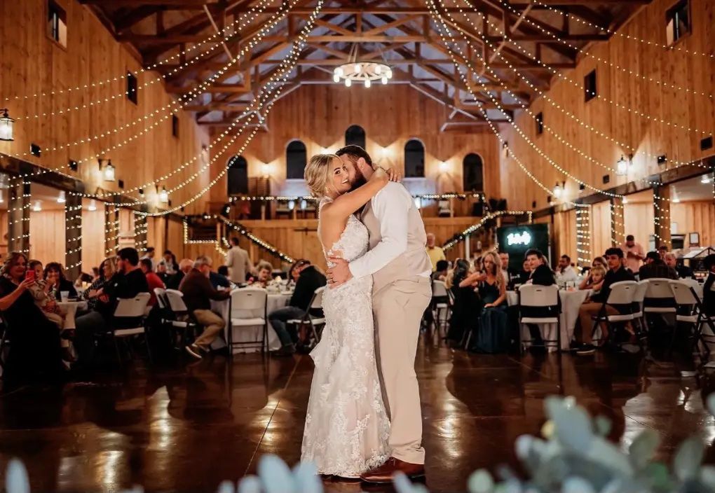 Couple dances at wedding reception in rustic barn, illuminated by string lights.