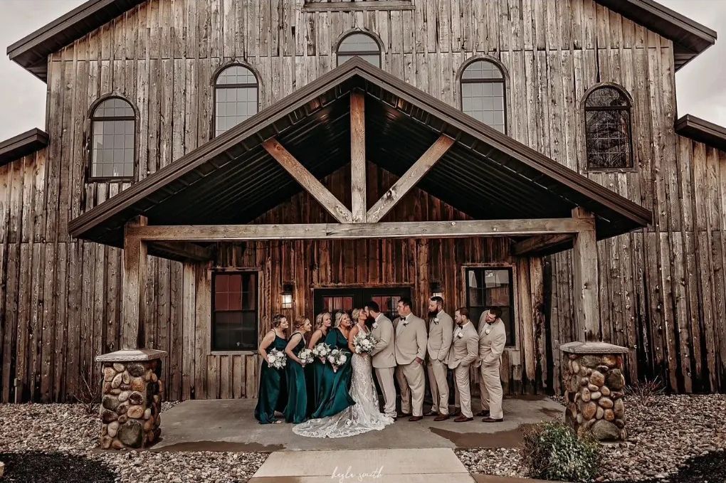 Wedding party posing in front of a weathered wooden building; bridesmaids in teal, groomsmen in tan, bride and groom in the center.
