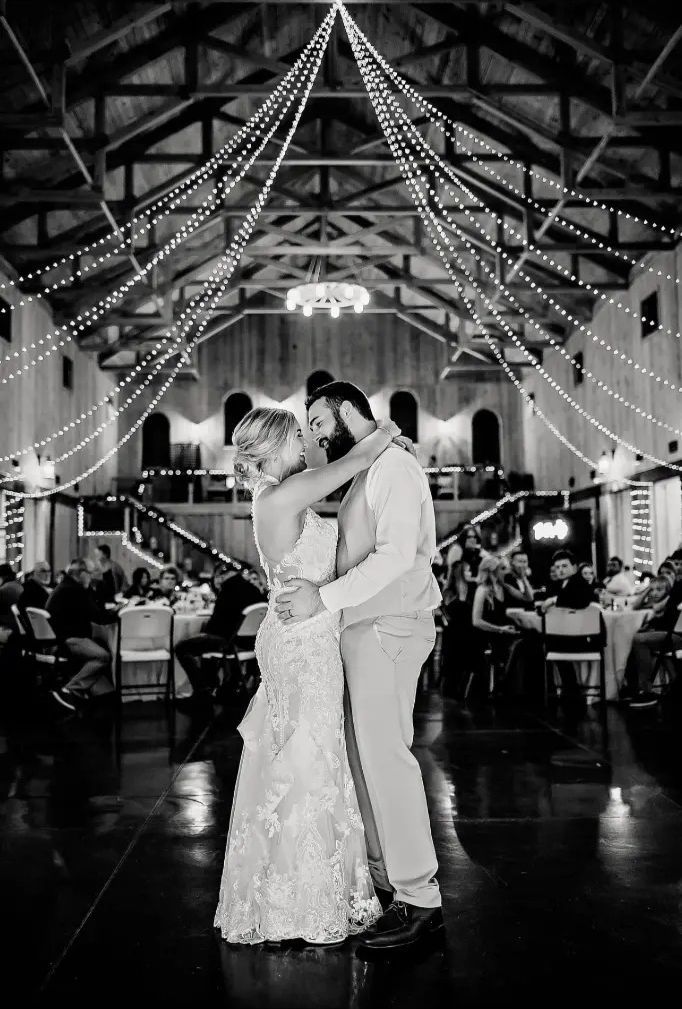 Bride and groom dance at reception in a rustic barn, string lights overhead.