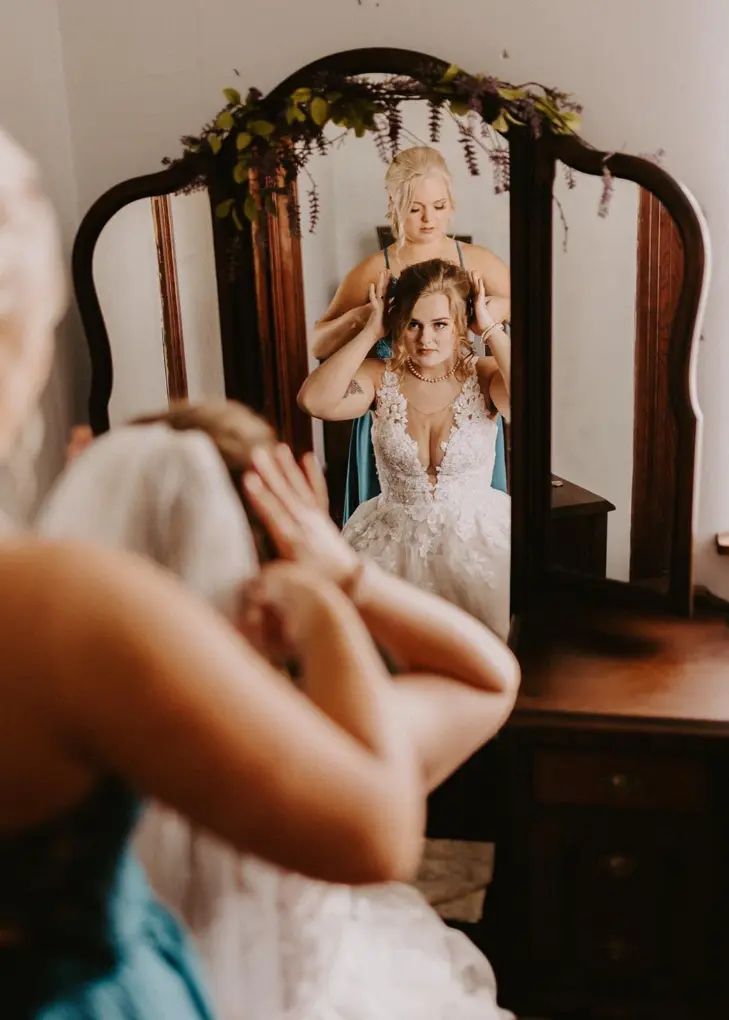 Bride having hair styled in front of a mirror with floral arch. Bridesmaids in the room.