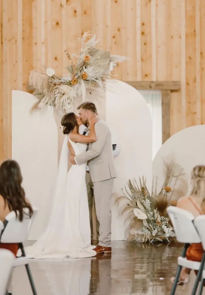 Newly married couple kissing at wedding ceremony, in front of a white backdrop and floral arrangement.