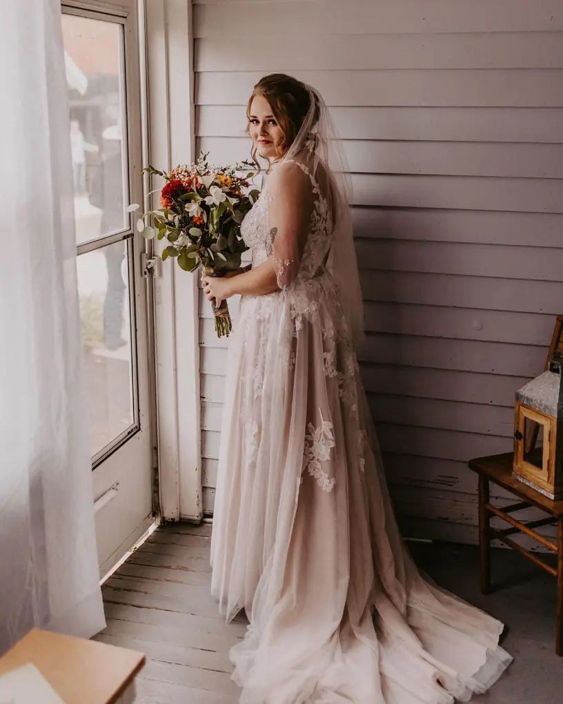 Bride in wedding dress, holding bouquet, poses by a window, light streaming in.