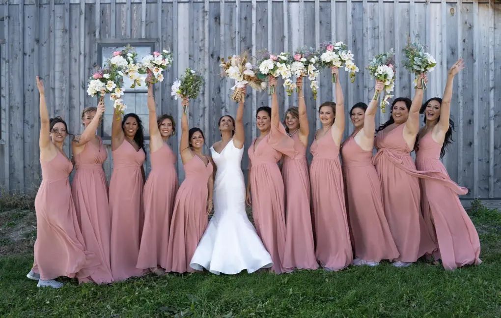 Bride in white dress with bridesmaids in pink dresses raising bouquets. Barn setting.