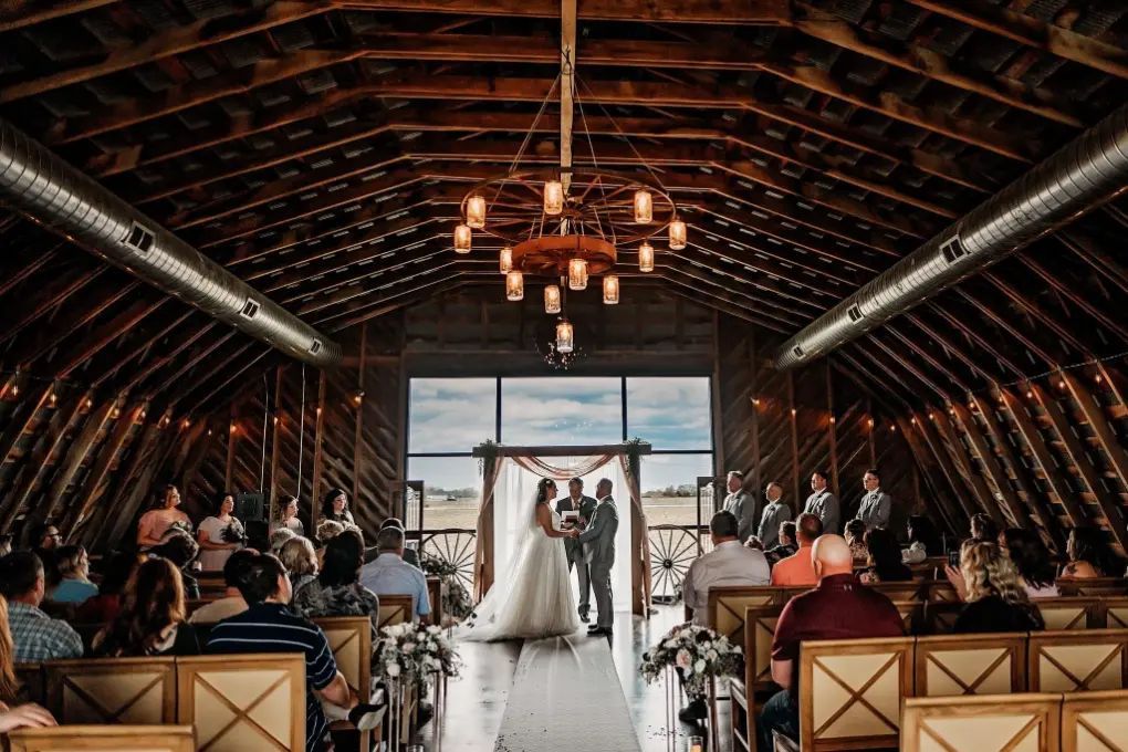 Wedding ceremony inside a barn with bride, groom, and guests. Wooden interior with chandelier and outdoor view.