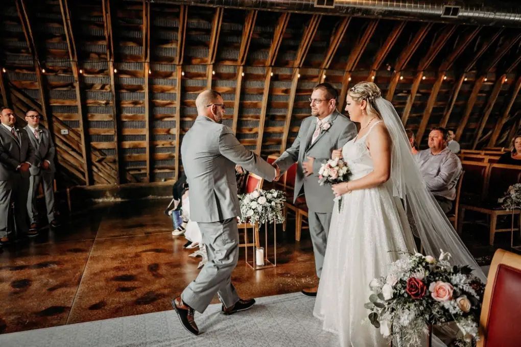Groom shakes hands with officiant as bride waits during a wedding ceremony in a rustic barn.