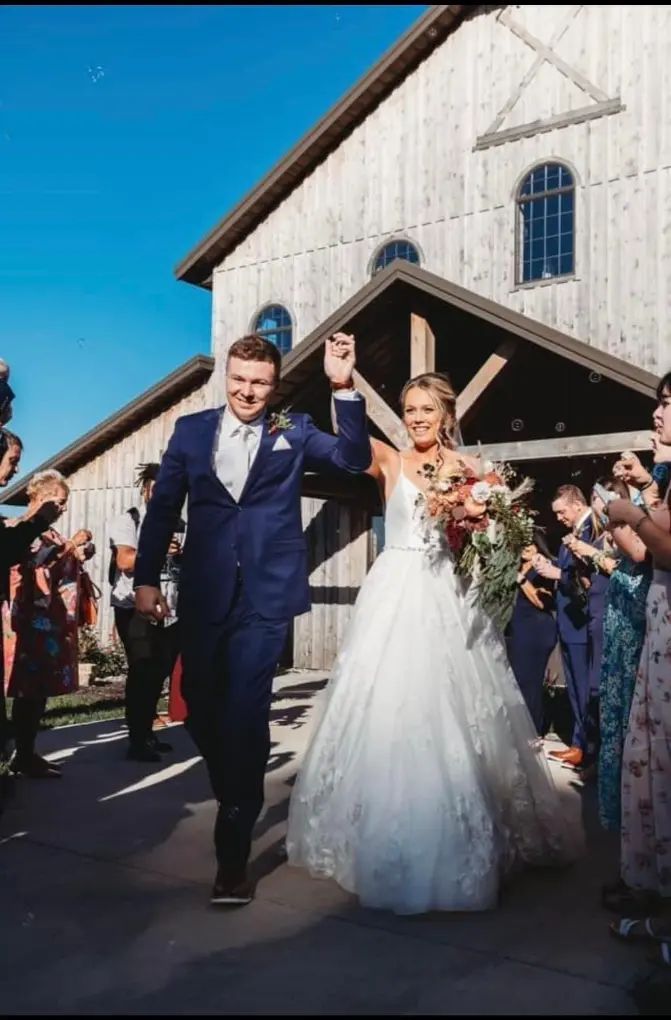 Newlyweds exiting a wooden barn, arms raised amidst guests holding sparklers, blue sky.