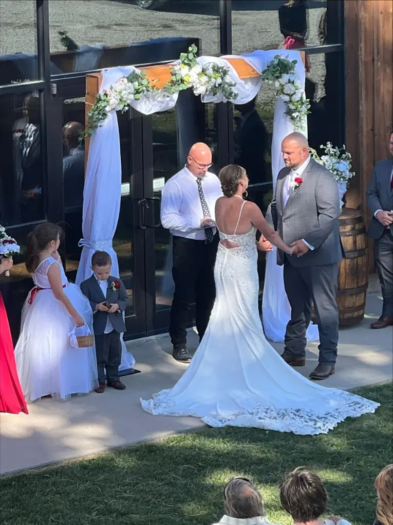 Wedding ceremony outdoors; couple exchanging vows under floral arch, officiant present.