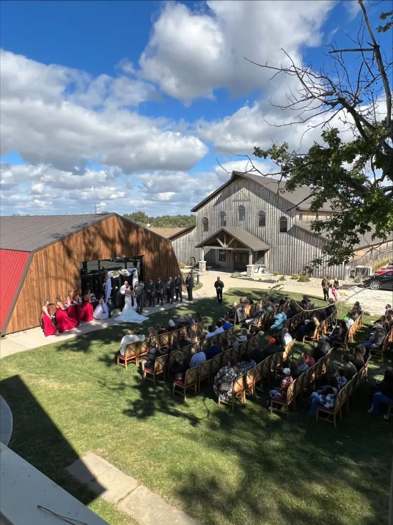 Wedding ceremony outdoors at a barn venue with bride, wedding party, and guests. Blue sky, sunny day.
