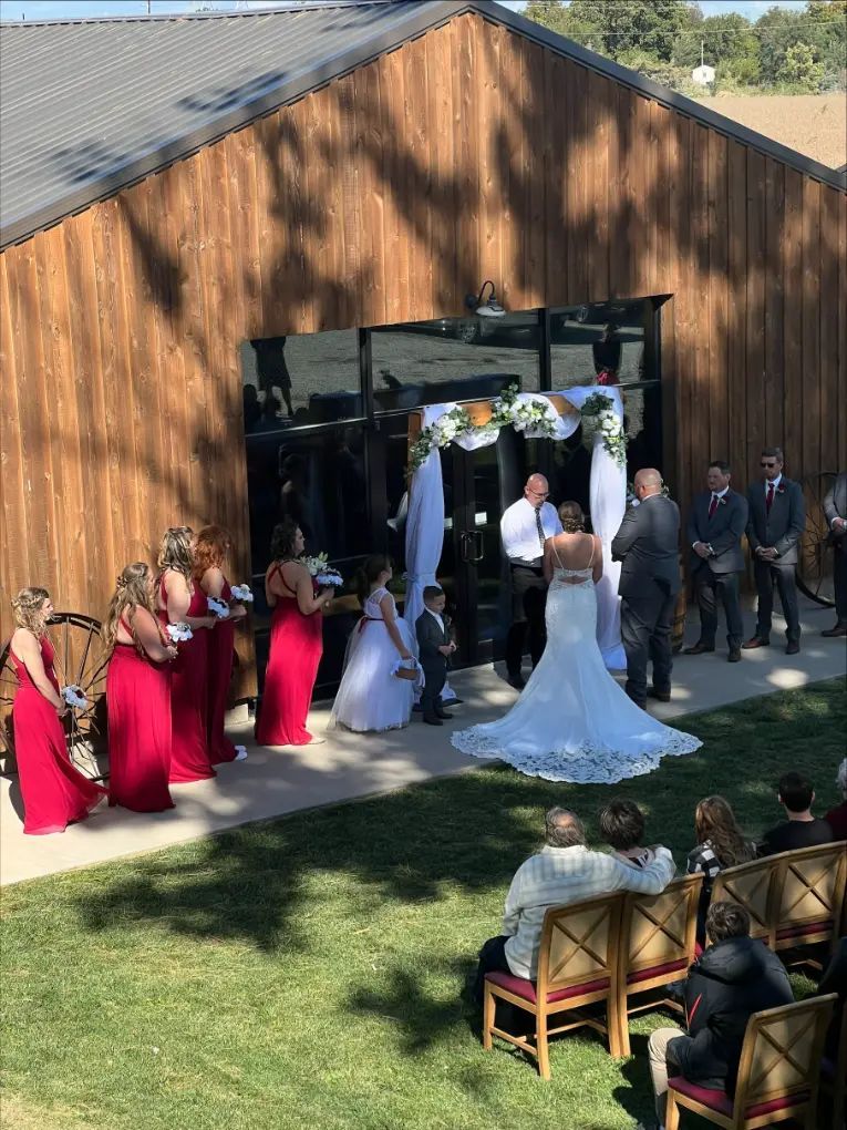 Wedding ceremony outside a wooden building. Bride in white dress, bridesmaids in red, guests seated.