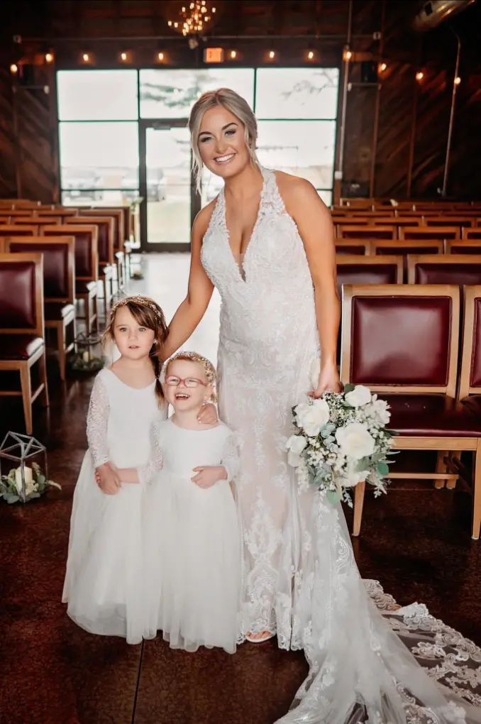 Bride in lace wedding dress with two flower girls, holding a bouquet, inside a ceremony venue.
