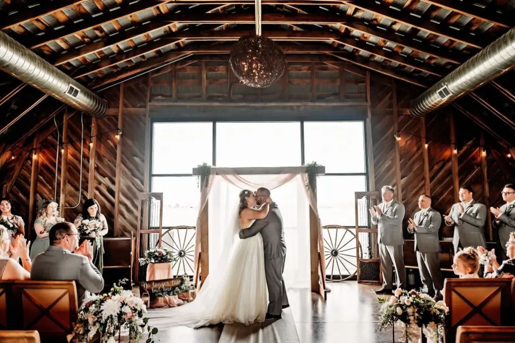Bride and groom kissing under a decorated archway inside a wooden building during their wedding ceremony. Guests watch.