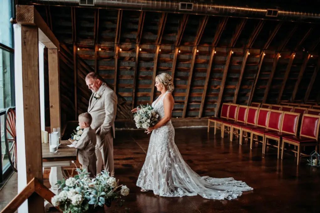 Bride with bouquet, groom, and child near altar in rustic barn with red chairs.