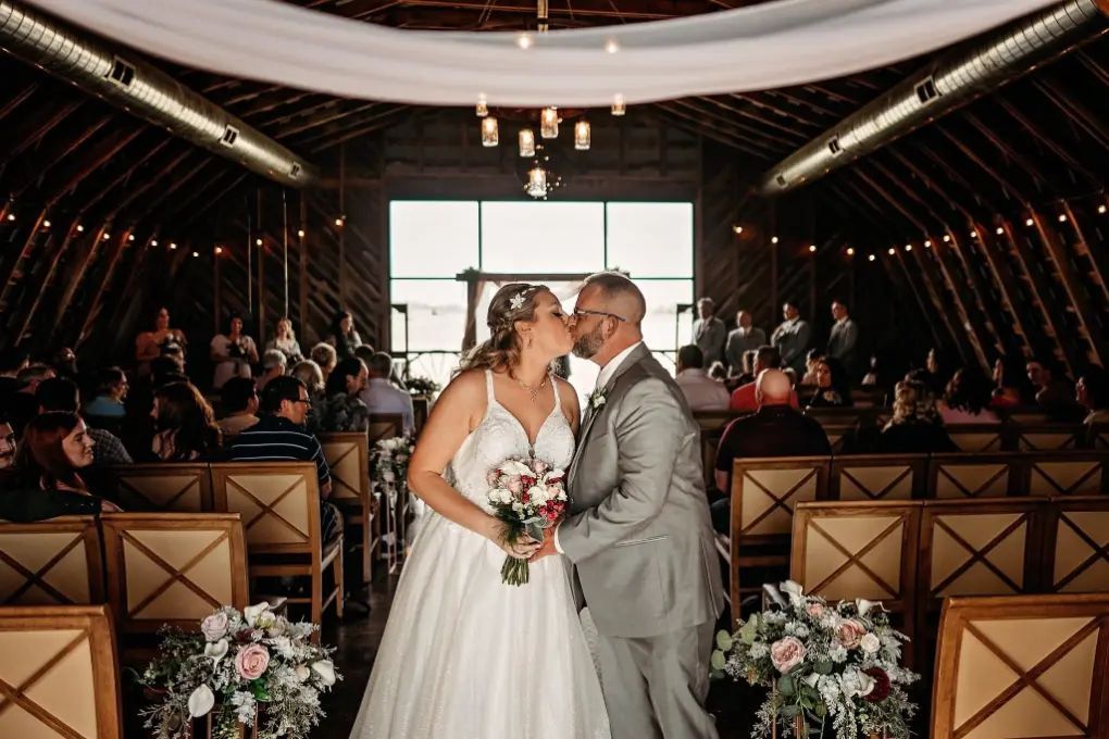 Bride and groom kiss at altar in a barn, surrounded by guests. Soft light and floral decorations.