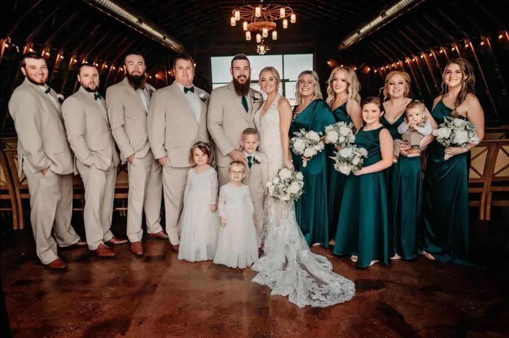 Wedding party posing indoors, featuring couple, groomsmen in tan, bridesmaids in teal.