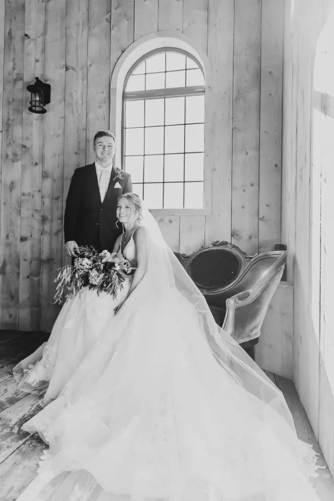 Bride and groom pose inside. Bride wears a veil and holds a bouquet, groom wears a suit. They stand near a window.