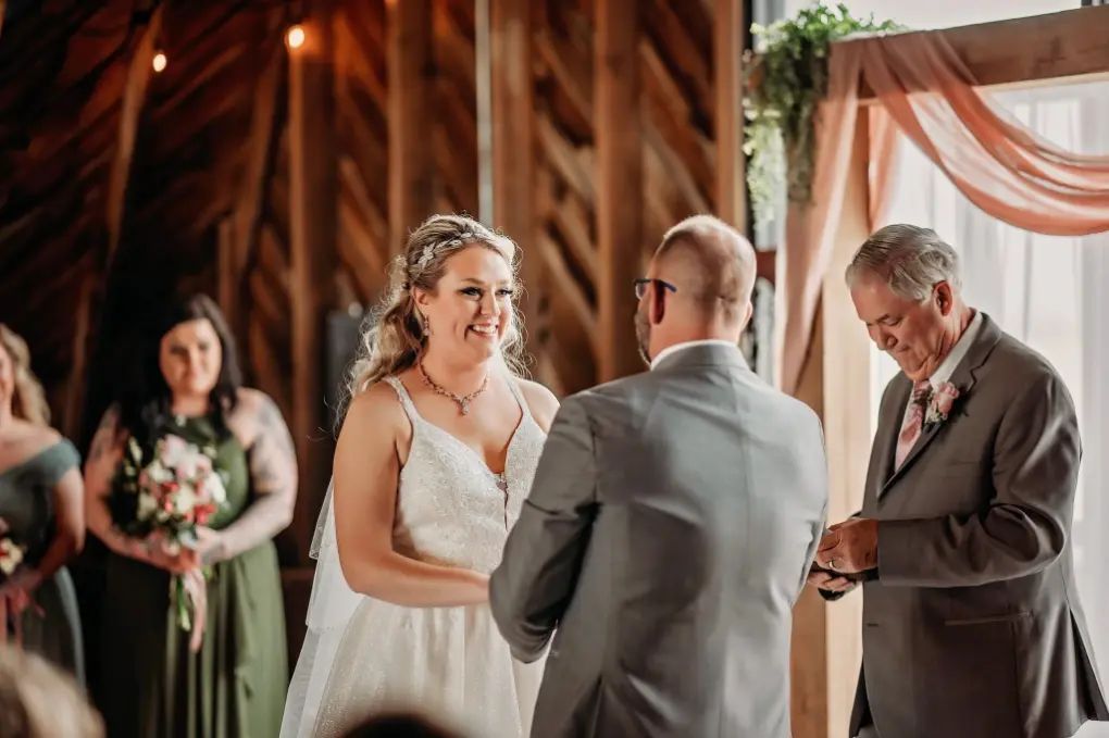 Bride and groom exchanging rings during a wedding ceremony. Bridesmaids in green dresses stand nearby. Wooden background.