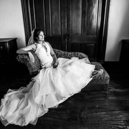 Bride in a white wedding dress sits on a chaise lounge in front of wooden doors.