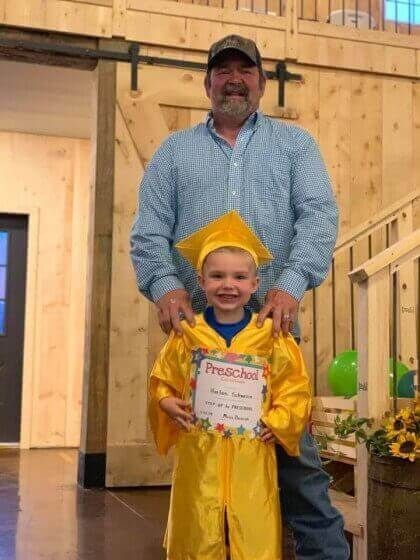 Man stands behind child in yellow graduation cap and gown, holding a diploma, smiling. Rustic interior setting.