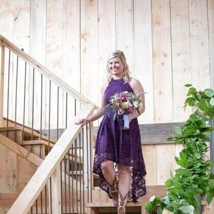 Woman in purple dress descending wooden stairs, holding flowers. Barn setting.