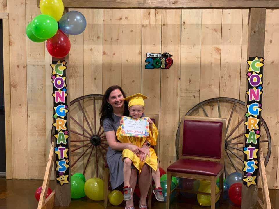 Woman and child in graduation attire with certificate, smiling, wooden backdrop, balloons.