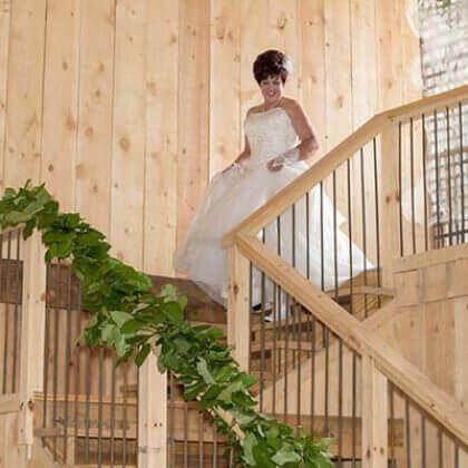 Bride in a white gown descending a wooden staircase decorated with greenery.