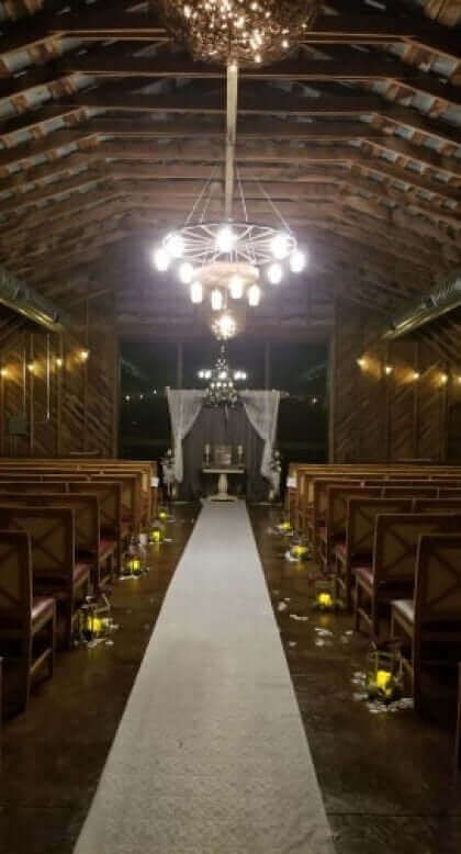 Wedding ceremony setup in rustic barn: aisle, seating, lit chandelier, lanterns, draped altar.