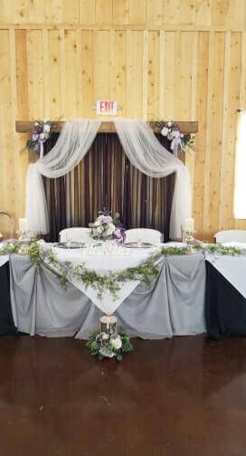 Wedding reception head table with gray and white draped fabric, greenery, and purple flowers.