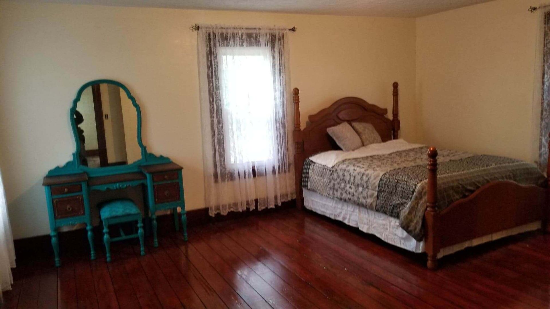 Bedroom with wooden floor, bed, and teal vanity with mirror. Window with sheer curtains.