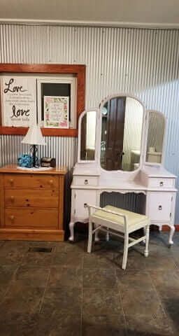 Pink vanity with tri-fold mirror and stool next to wooden chest and framed art on corrugated metal wall.