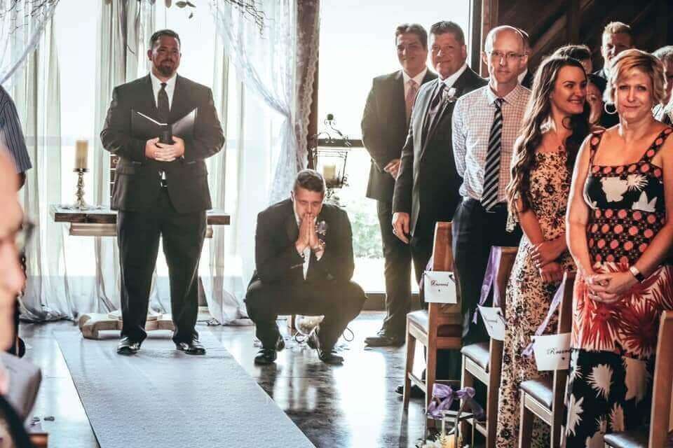 Man in suit squatted on aisle, appearing emotional; guests and officiant wait at a wedding.