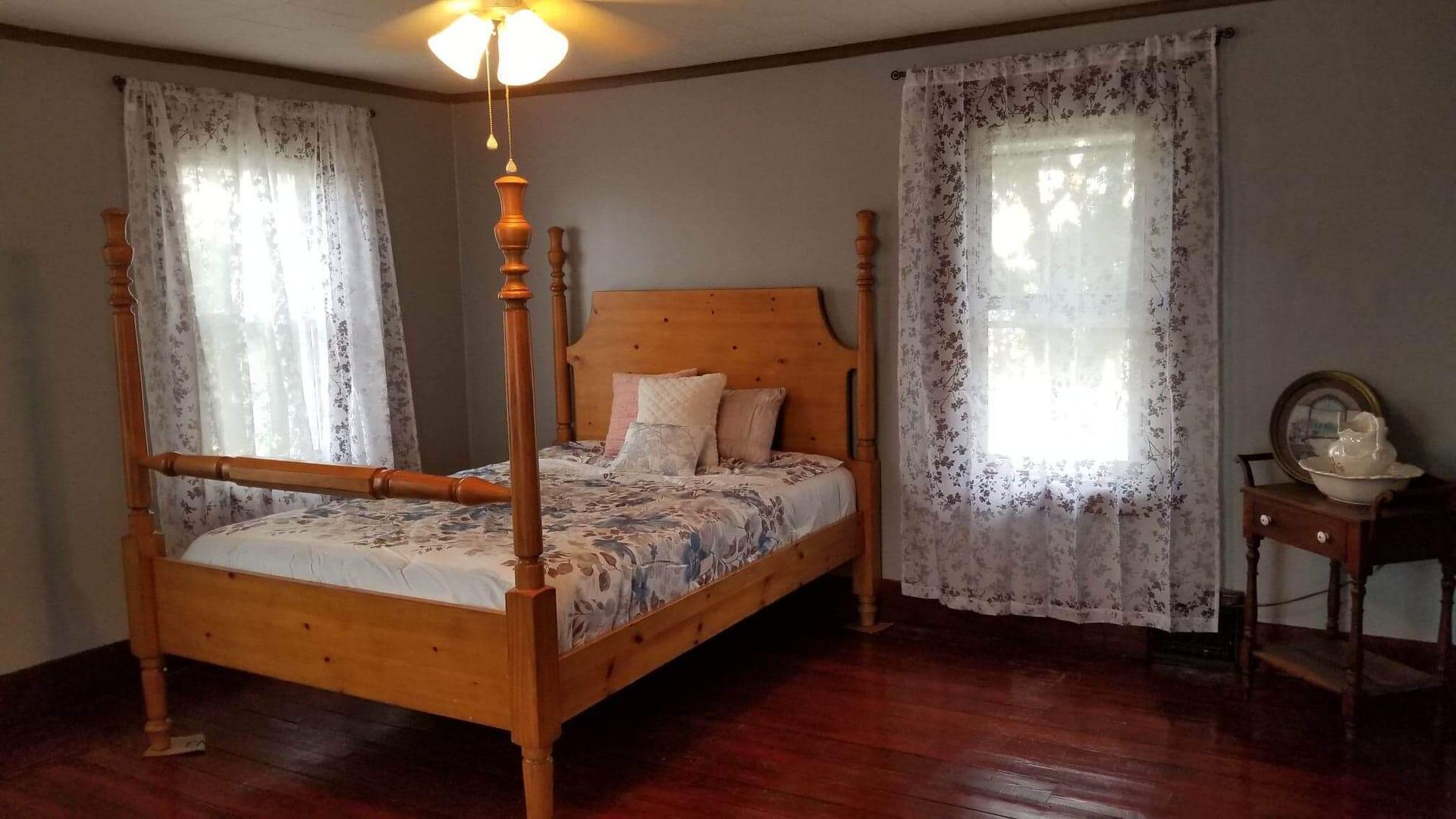 Bedroom with wooden four-poster bed, floral bedding, lace curtains, and small side table.