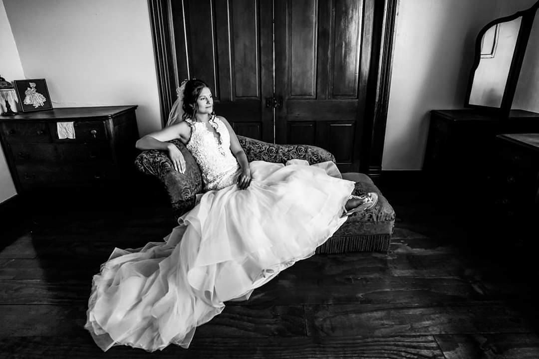 Bride in wedding dress, seated on a chaise lounge, against dark wooden doors.