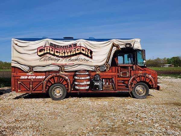 Food truck designed as an old-fashioned chuckwagon, red and brown with canvas top and barrels, parked on gravel.