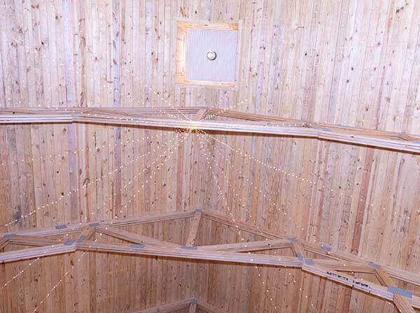 Interior wooden ceiling, circular shape. Strings with small lights hang from the center. Square opening at the top with a light fixture.