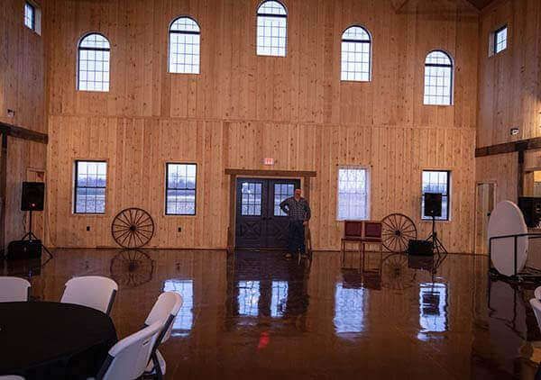 Spacious wooden interior with windows, glossy floor. Person stands near doorway. Tables and chairs in foreground.