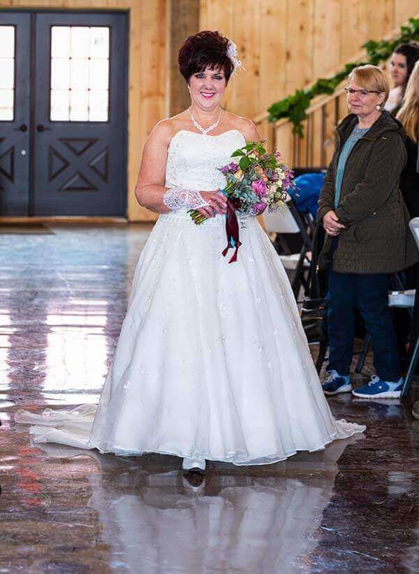 Bride in a white strapless gown holding flowers, walking toward the camera at a wedding.