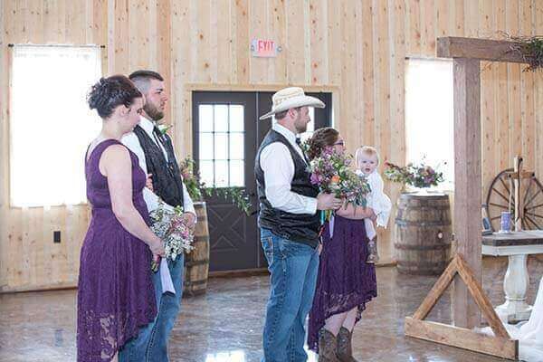 Wedding ceremony inside a wooden building. People stand near an archway. Bridesmaids in purple dresses. Groomsmen wear vests and jeans.