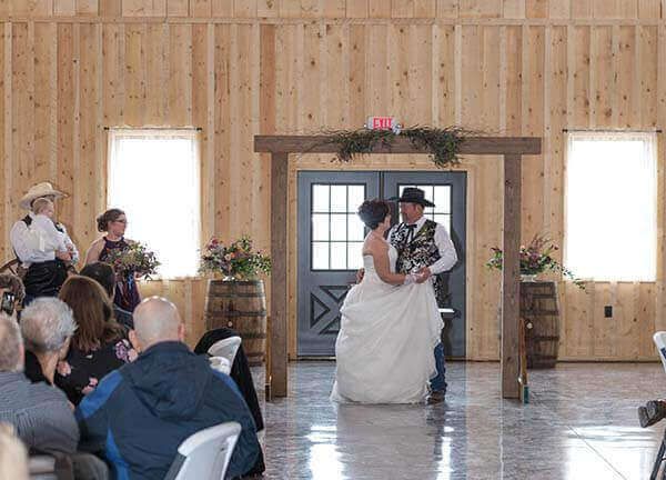 Couple dances at wedding ceremony in wooden barn, guests watch, arch with flowers.