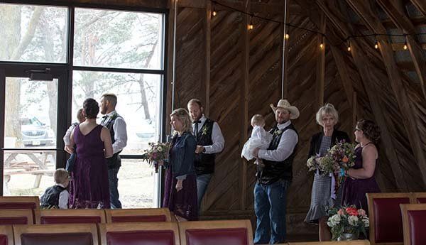 Wedding party stands inside rustic building with large window, holding flowers.