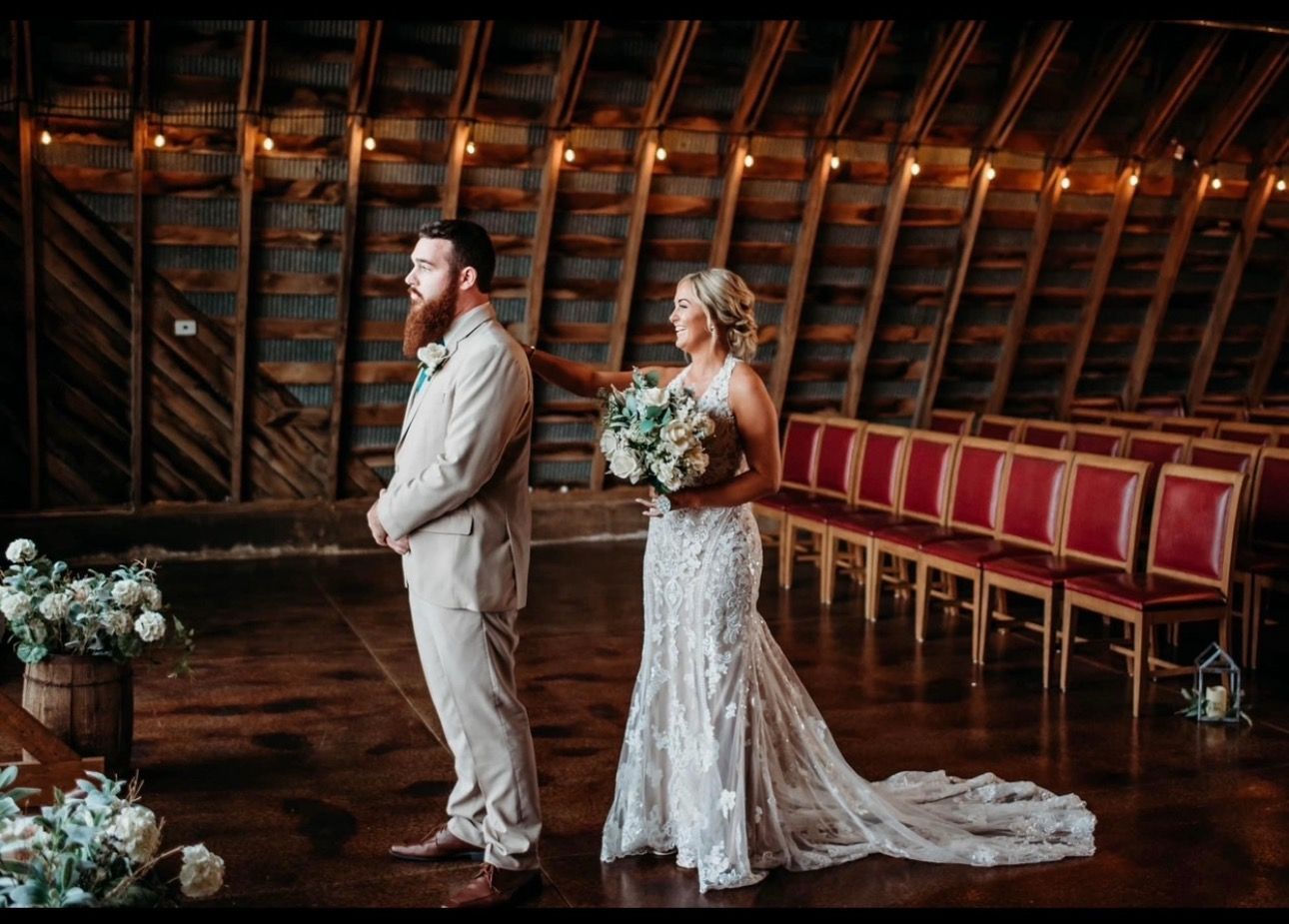 Bride approaching groom at wedding ceremony in rustic barn. Bride in white dress, groom in tan suit.