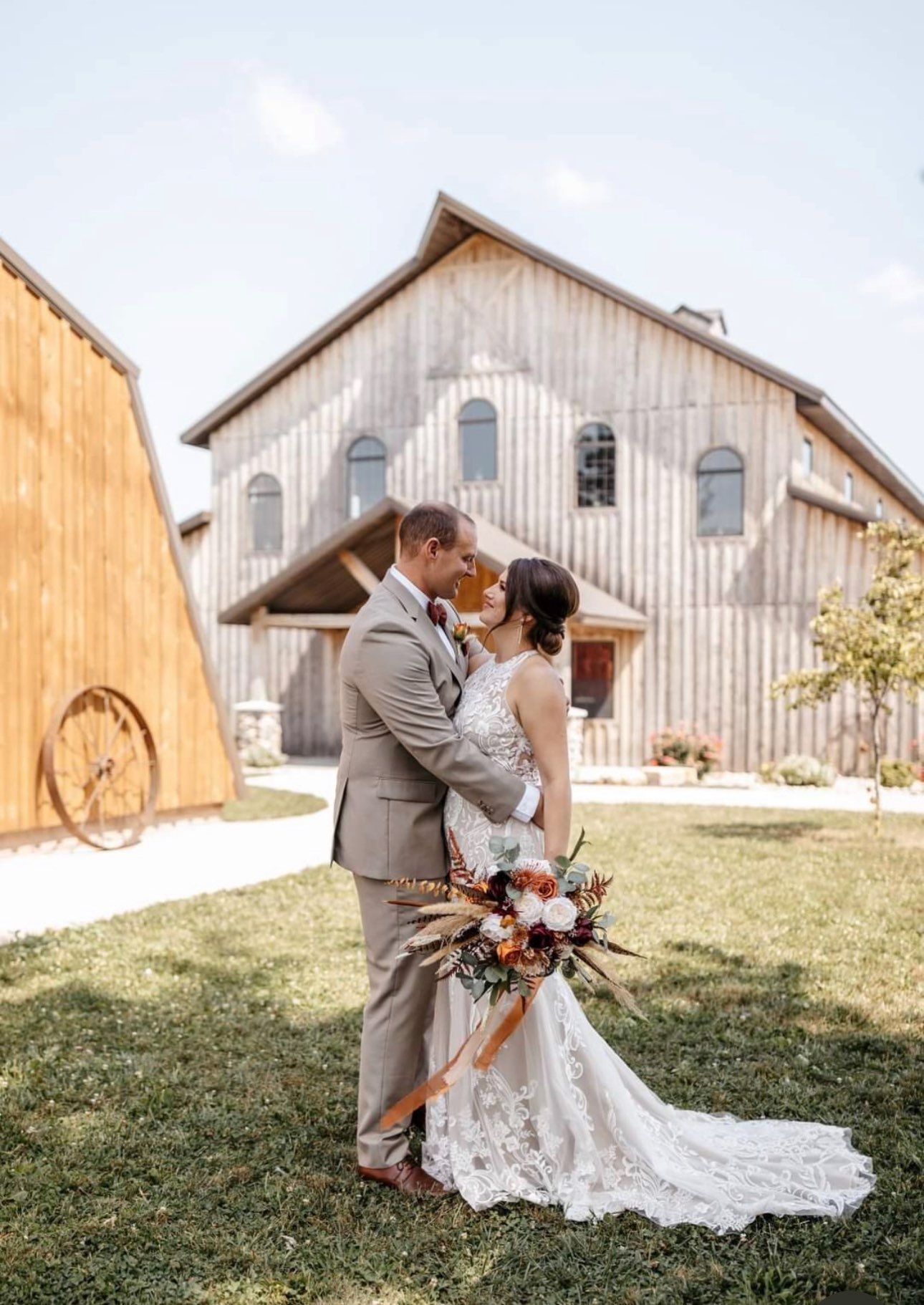 Wedding couple embraces in front of a barn; bride holds bouquet.