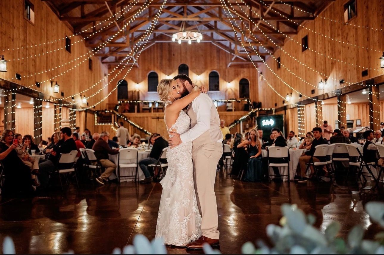 Newlyweds share a kiss during their first dance at a warmly lit wedding reception in a barn.