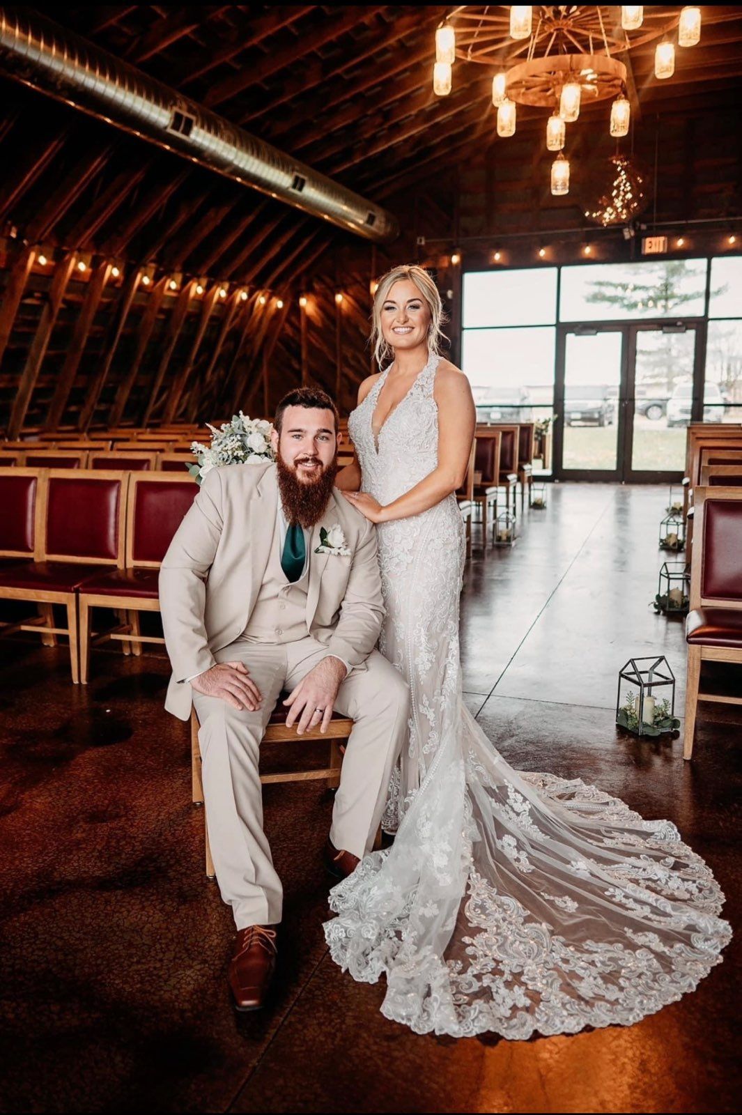 Bride and groom in wedding attire posing inside barn. She wears a white gown, he wears a tan suit.