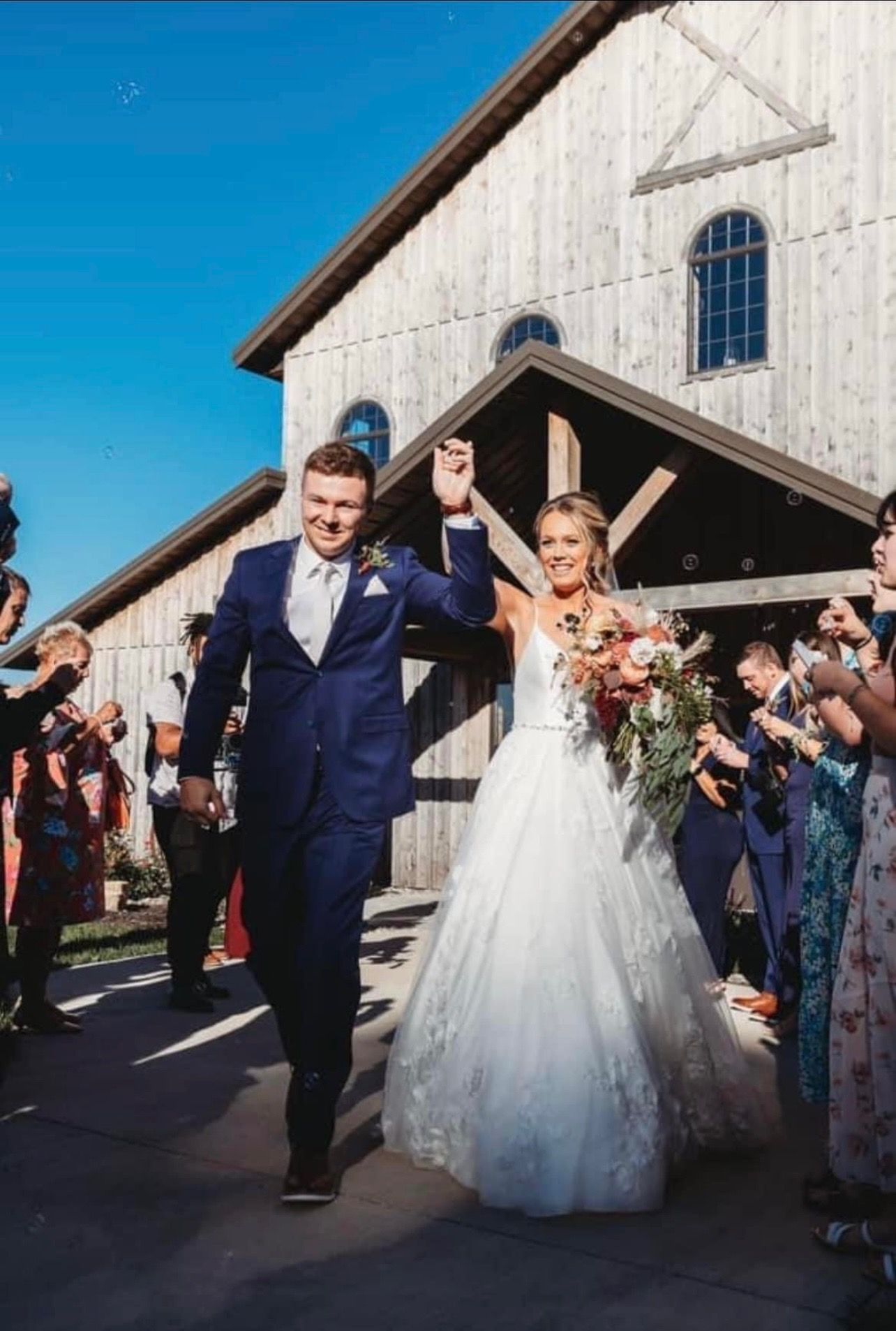 Newlyweds exit venue waving, showered with confetti. Groom in blue suit, bride in white gown, sunny day.