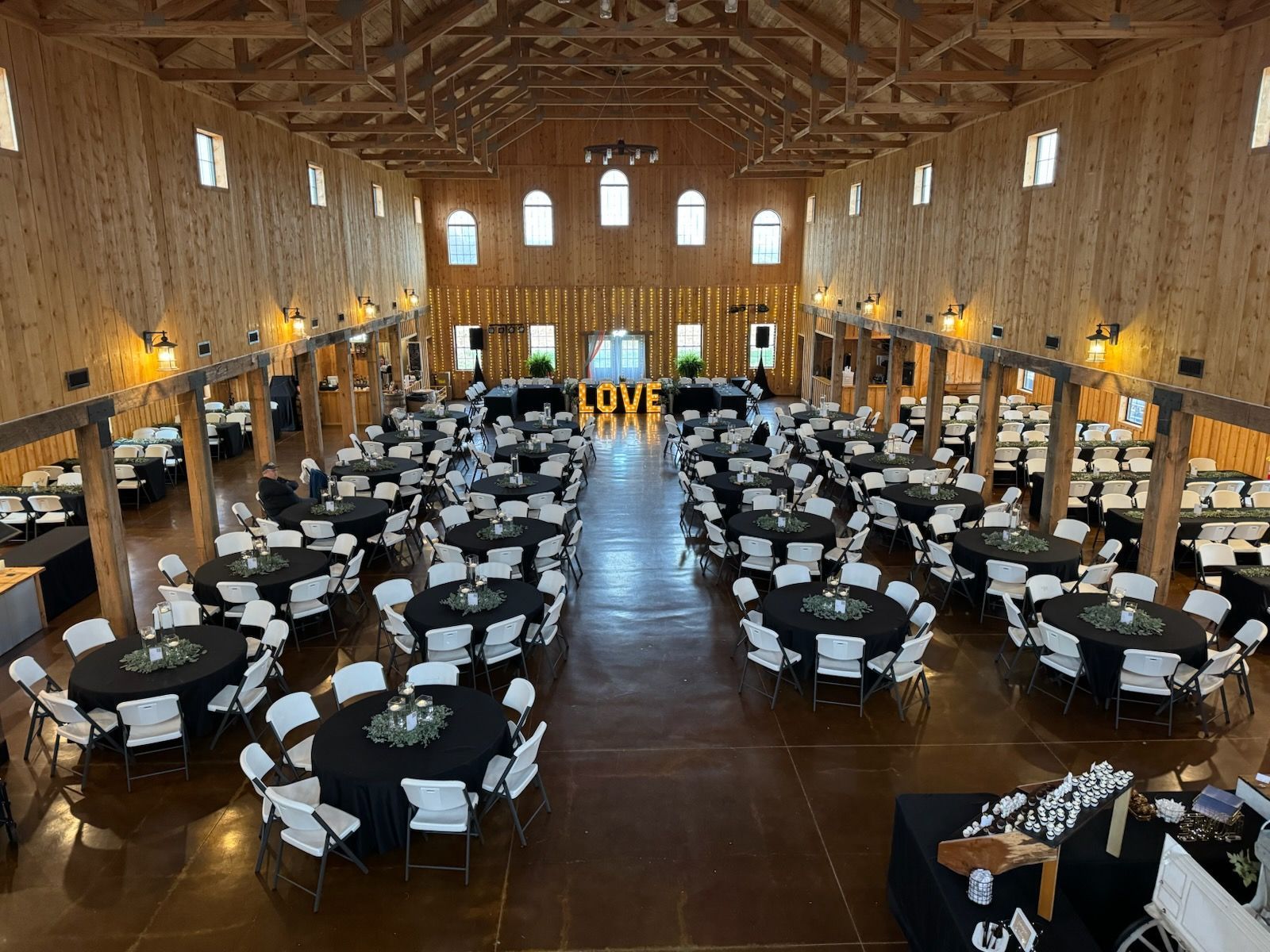 Large event hall with round tables covered in black linens, white chairs, and wooden beams.