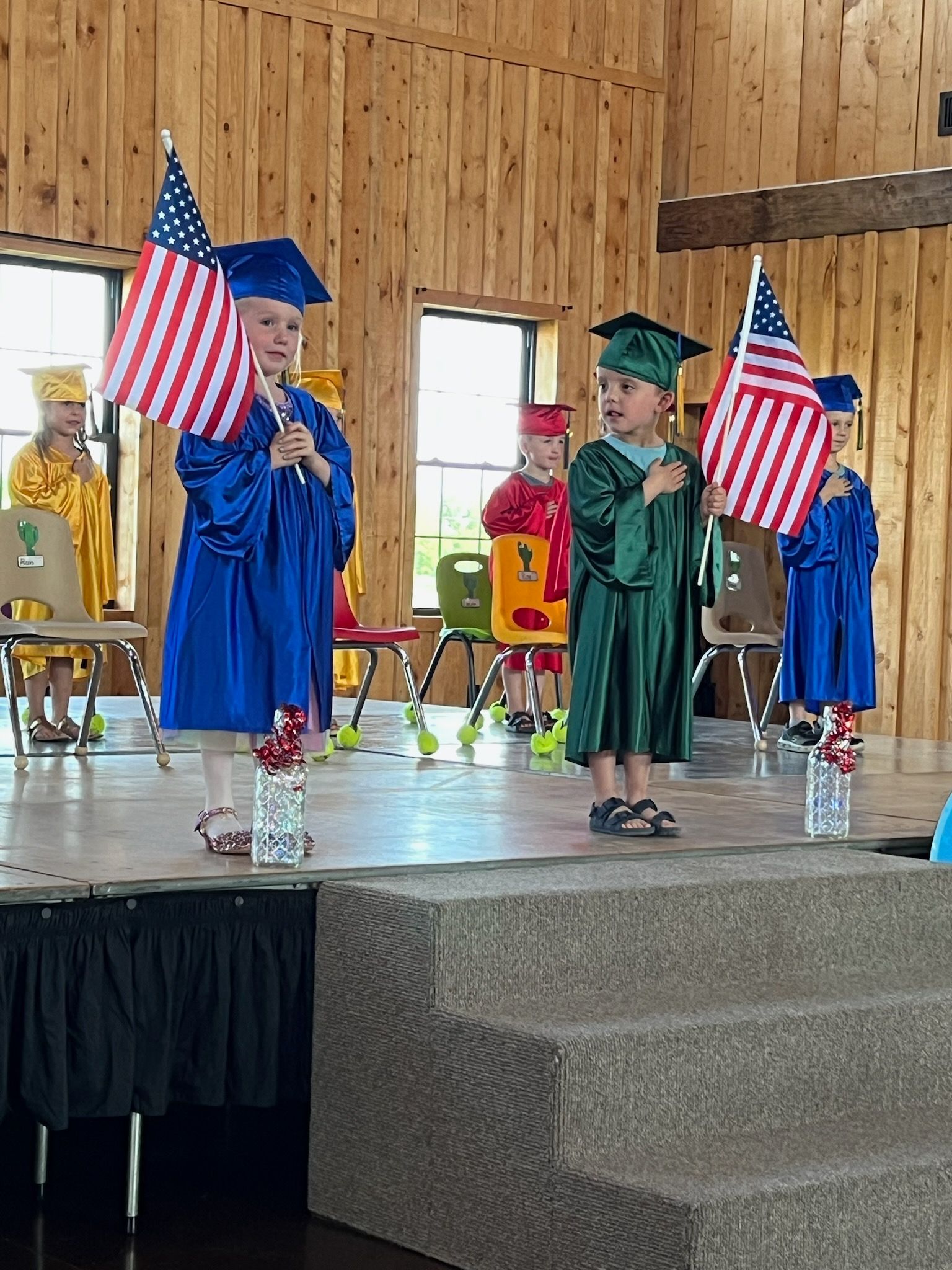 Children in graduation gowns holding American flags on a stage indoors.