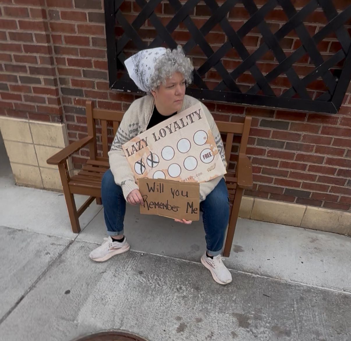 Old woman dressed as a paper punch card sitting on a bench holding a cardboard sign that reads “Will you remember me,” symbolizing an outdated loyalty program.